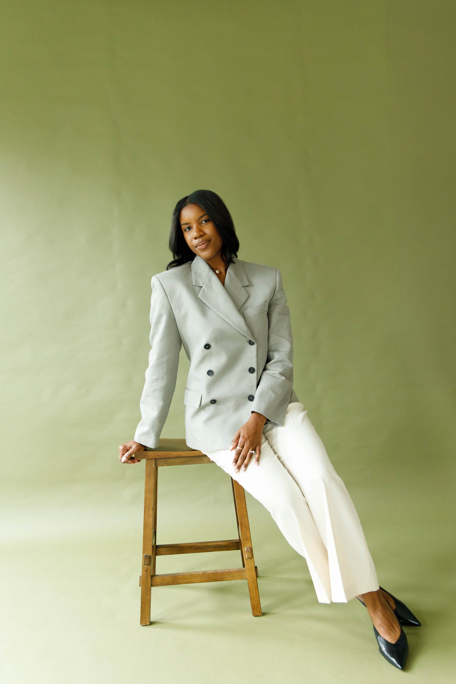 A woman sits on a wooden stool wearing a light gray double-breasted blazer and white wide-leg trousers, posed against a solid sage green studio backdrop.