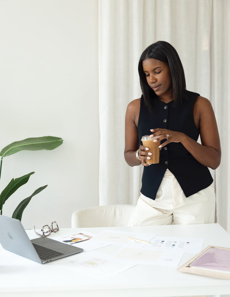 Woman sitting at a desk with a laptop, holding a coffee cup in a bright room.